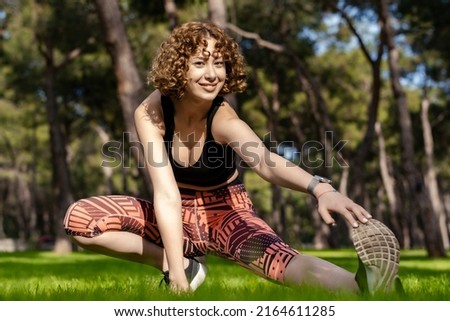 Cute caucasian woman wearing black sports bra standing on city park, outdoors stretching leg muscles, doing side lunges in skandasana pose. Healthy lifestyle concept.
