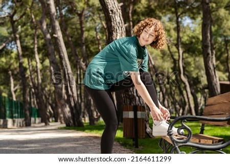 Cheerful redhead woman wearing green t-shirt standing on city park, outdoor tying running shoes leaning on park bench getting ready for run. Jogging girl exercise motivation health and fitness.