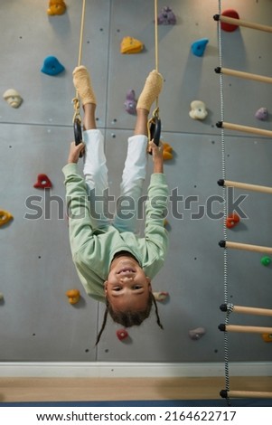 Full length portrait of active black child hanging upside down on sports rings at home and having fun