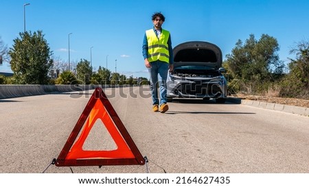 Young man preparing a red triangle to warn other road users, car breakdown or engine failure stop at countryside street. Male driver standing near a broken car with open up hood