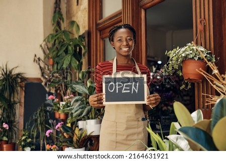 Happy African American flower shop owner holding open sign and looking at camera.