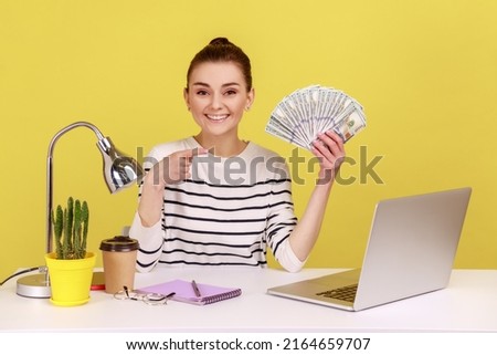 Positive young woman pointing finger at hundred dollar bills, holding sitting at workplace with laptop, high salary, bonuses and perks. Indoor studio studio shot isolated on yellow background. Royalty-Free Stock Photo #2164659707