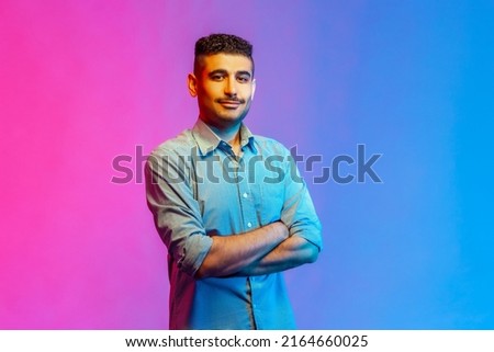 Portrait of calm smiling man in shirt standing with crossed arms, looking at camera with confident facial expression. Indoor studio shot isolated on colorful neon light background.