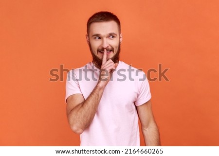 Portrait of positive bearded man showing hush with finger on his lips gesture, shushing, asking to keep silence, wearing pink T-shirt. Indoor studio shot isolated on orange background.