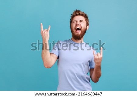 Portrait of bearded man showing rock and roll gesture heavy metal sign, enjoys favorite music on party, has fun squints face exclaims from joy. Indoor studio shot isolated on blue background.