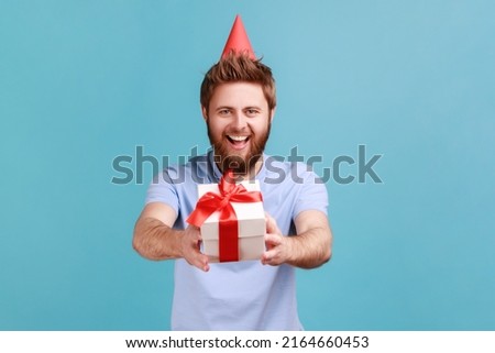 Portrait of man in party cone giving birthday gift and smiling, sharing present, donating and offering surprise in box, congratulation on anniversary. Indoor studio shot isolated on blue background.