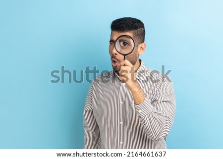 Serious bearded man standing, holding magnifying glass and looking at camera with big zoom eye, verifying authenticity, wearing striped shirt. Indoor studio shot isolated on blue background.