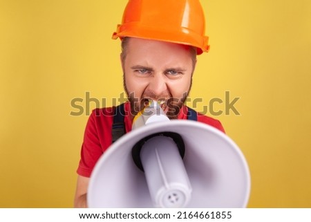 Portrait of angry aggressive worker man wearing uniform and protective helmet taking selfie, point of view of photo, screaming in megaphone. Indoor studio shot isolated on yellow background.
