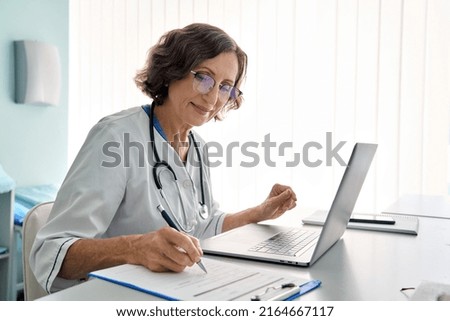 Senior middle aged female medical worker in modern clinic wearing white doctor's coat having videocall using laptop writing health personal data, consulting remotely. Telemedicine healthcare concept. Royalty-Free Stock Photo #2164667117