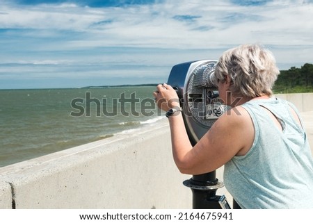 An elderly woman looking through the tourist telescope turned towards infinity. Seascape of the blue Atlantic ocean. Coin-operated binoculars for landscape exploration