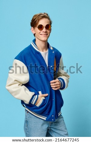 a happy, handsome, fashionable student in a blue bomber jacket is standing in sunglasses and with a bright backpack on his back. Vertical studio photography Royalty-Free Stock Photo #2164677245