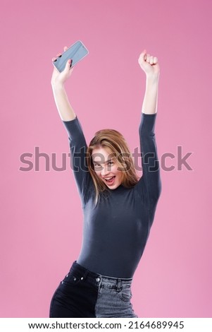 A young and beautiful brunette posing on a pink background whit a joyful expression, holding her hands and phone in the air Royalty-Free Stock Photo #2164689945