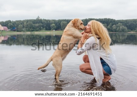 Young beautiful woman with blond curly hair huging with her labrador retriever dog on the river shore
