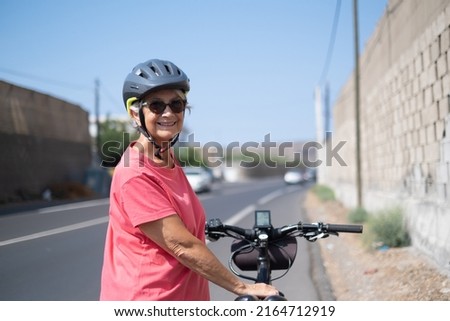 Cheerful caucasian senior woman, wearing protective helmet, running on her electric bicycle in the sunny road. Healthy lifestyle and sustainable mobility concept
