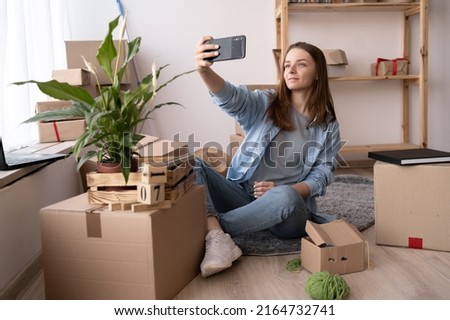 happy brunette girl taking selfie with smartphone and sitting on floor among cardboard boxes at home, new home, technology, people, renovation and relocation concept