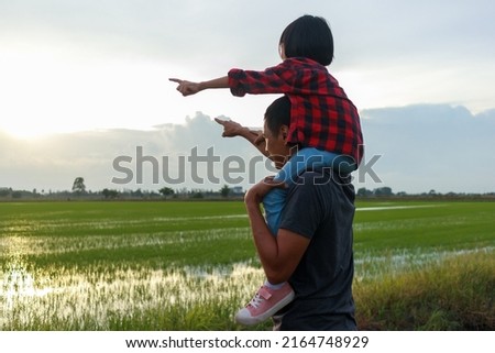 Picture of daughter riding father's neck They both stood in the fields. having growing green rice seedlings And they're pointing to the last light of day. they are happily.  green background.