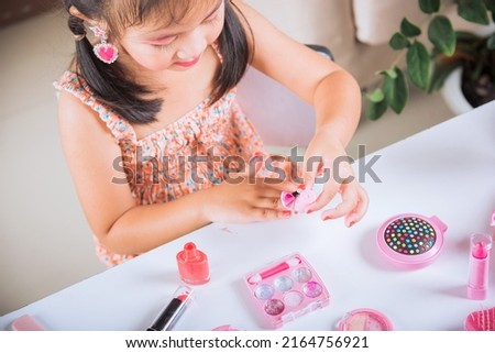 Asian adorable funny little girl making makeup dips brush into bottle to paints nails polish red nail varnish herself, Learning activity to be woman, happy kid is beautiful make up with cosmetics toy Royalty-Free Stock Photo #2164756921