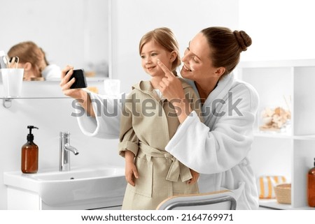 beauty, family and people concept - happy smiling mother and daughter with moisturizer on their faces taking selfie with smartphone in bathroom