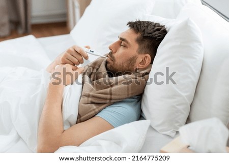 people, health and fever concept - sick man in scarf measuring temperature by thermometer lying in bed at home Royalty-Free Stock Photo #2164775209