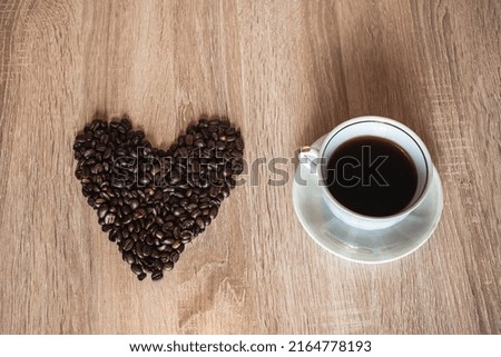 A cup of hot aromatic coffee in a saucer, against the background of scattered coffee beans in the form of a heart, on a wooden light brown table.