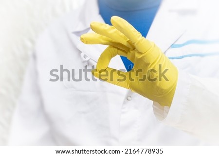 A doctor in a medical gown and protective gloves shows an OK gesture. The face is not visible.