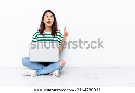 Young Vietnamese woman with a laptop sitting on the floor isolated on white wall pointing up and surprised
