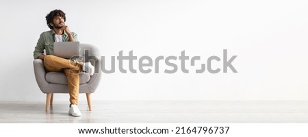 Pensive millennial indian guy in casual sitting in armchair with computer on lap, touching chin and looking at copy space over white studio background, panorama, business opportunities concept