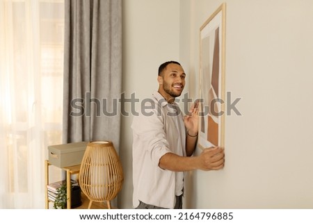 Smiling young black man putting picture frame, hanging painting on wall, empty space. African American male interior designer decorating new modern stylish apartment. Home interior and domestic decor Royalty-Free Stock Photo #2164796885