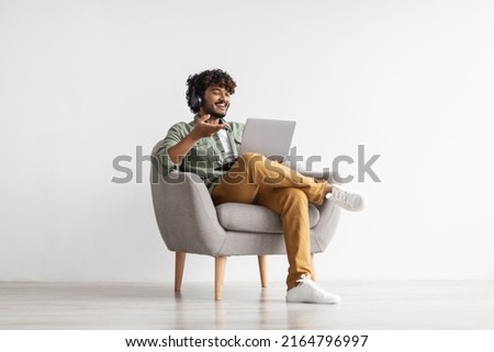 Positive handsome young indian guy sitting in armchair over white studio background, using laptop and headset, having video call with friend or lover, copy space. Modern technologies and communication Royalty-Free Stock Photo #2164796997