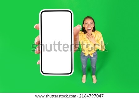 Excited Female Showing Big Cellphone With Empty Screen Smiling To Camera Standing In Studio Over Green Background. Lady Advertising Mobile Offer Or Application. High Angle, Mockup