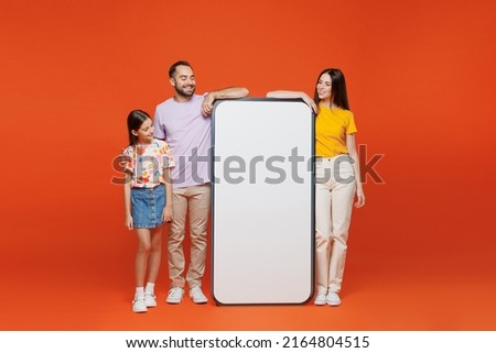 Full body young parents mom dad with child kid daughter teen girl wear basic t-shirts big blank screen mobile cell phone with workspace mockup area isolated on yellow background. Family day concept.