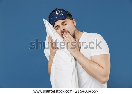 Close up young man in pajama jam sleep mask rest relax at home hold hug pillow lying in bedroom yawning cover mouth with hand isolated on dark blue background studio Good mood night bedtime concept.