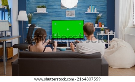 Couple of gamers spending free time playing fast paced console action game on green screen competing against each other. Young woman and boyfriend gaming online having fun on chroma key display.