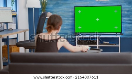 Static tripod shot of young woman watching movie on tv with green screen and eating popcorn while sitting on couch. Girl looking at favourite television show on chroma key display in home living room.