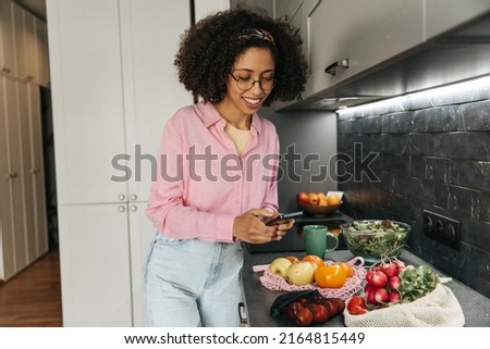 Cheerful young african woman taking pictures of her vegetable purchases in kitchen. Brunette wears glasses, shirt and jeans. Technology concept
