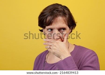 Attractive mature woman in purple blouse on a yellow background with hand over mouth looking away to camera right