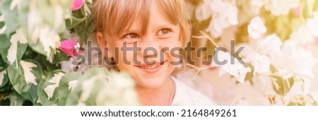 portrait of face candid little happy smiling five year old blonde kid boy with green eyes in pink and white flower plants in nature. children have fun summer holidays. banner. flare