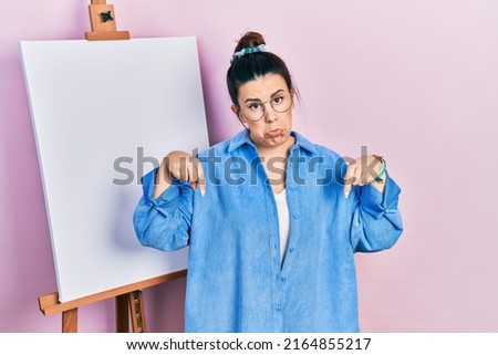 Young hispanic woman standing by painter easel stand pointing down looking sad and upset, indicating direction with fingers, unhappy and depressed. 