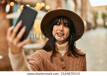 Brunette woman wearing winter hat taking a selfie picture with smartphone outdoors at the city