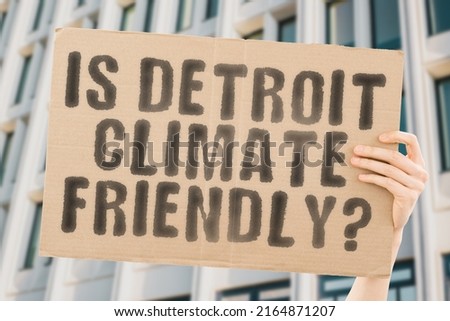 The question " Is Detroit climate-friendly? " is on a banner in men's hands with blurred background. Support. Team. Activist. Urban. Sunset. Carbon. Ecology. Energy. New. Clean. Warming. Waste