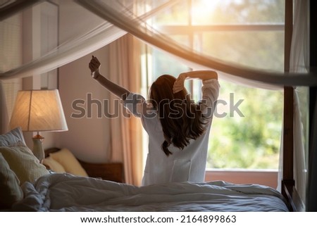 Back view of a blurry soft image of a woman with long hair is stretching relaxing on a sunny morning in a cozy room at a Resort hotel with green nature outside. Weekend, travel, and Holliday concept.