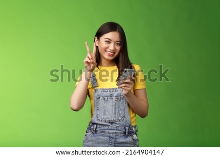 Cute tender asian woman hold smartphone, show victory peace sign, look camera delighted, carefree pose near green background, win online giveaway, triumphing joyfully beat score game