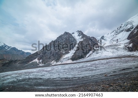Dramatic landscape with glacier tongue and icefall on large snow mountain range with sharp rocky pinnacle under gray cloudy sky. Long glacier in high altitude. Gloomy scenery in mountains in overcast.
