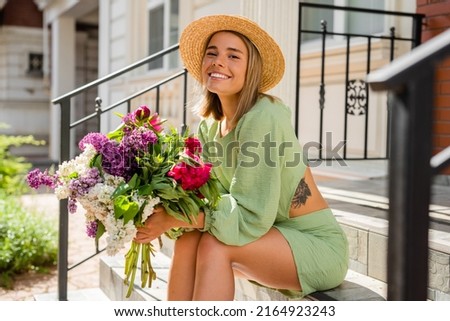 beautiful young woman in summer style outfit smiling happy walking with flowers in city street wearing straw hat fashion trend, sitting on stairs Royalty-Free Stock Photo #2164923243