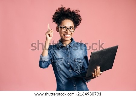 Overjoyed happy multiracial woman pointing finger up and smiling toothy while holding laptop. Having idea, business plan, startup concept. Indoor studio shot isolated on pink background Royalty-Free Stock Photo #2164930791