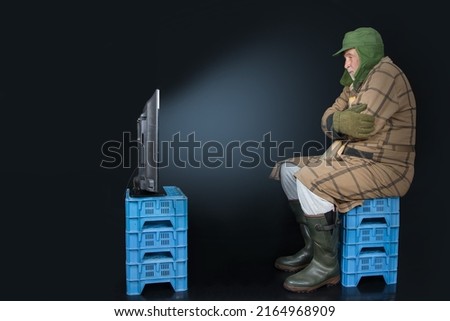 Freeze for Peace. TV evening autumn 2022 in Germany? A man, dressed warmly, is sitting on plastic boxes in front of the TV. He burned his furniture, oil and gas are too expensive because of war Royalty-Free Stock Photo #2164968909