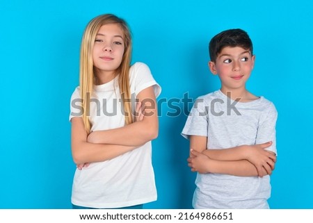 Waist up shot of  self confident two kids boy and girl standing over blue background has broad smile, crosses arms, happy to meet with colleagues.