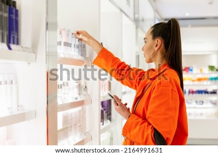 Woman chooses cosmetics in the store. A young beautiful brunette in a bright orange jacket holds a bottle of cream in her hands. Side view. Personal care and modern cosmetology.