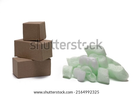Close up of a stack of small cardboard shipping boxes next to a pile of large soft green white packing chips flakes to protect fragile goods during shipping. Royalty-Free Stock Photo #2164992325