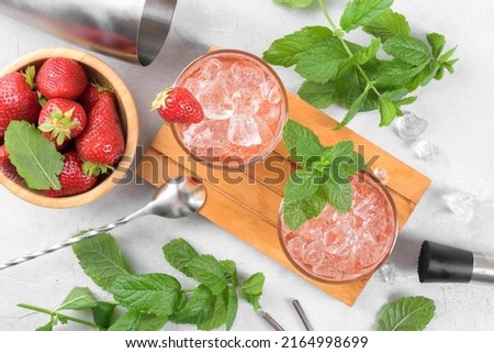 Cold summer strawberry cocktail mojito, margarita, daiquiri. Two glasses with fresh strawberry soda drink, ice cubes, mint and bartender tools on light background. Iced strawberry lemonade, top view